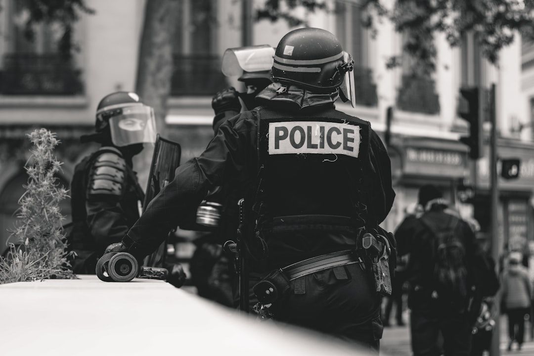 The image depicts police officers in tactical gear during a public order situation. One officer is prominently featured from behind, wearing a helmet and face shield, with 'POLICE' clearly visible on their uniform.