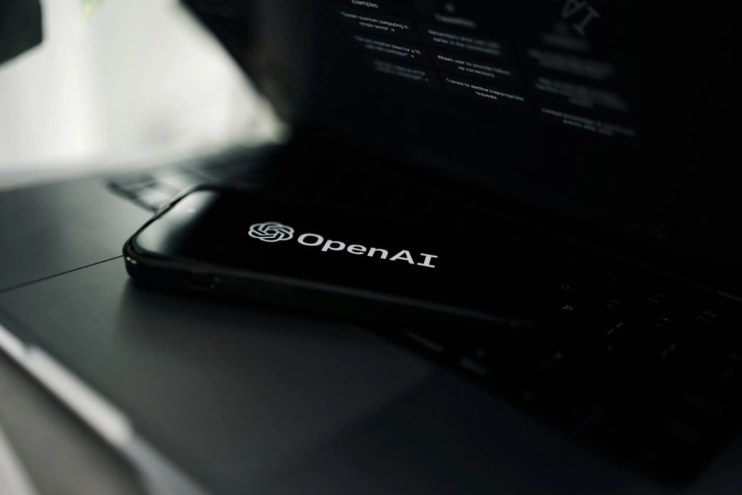 The image shows a smartphone displaying the OpenAI logo placed on a laptop keyboard. The background is muted, emphasizing the contrast between the black phone and the metallic laptop surface. — Credit: Photo by Zac Wolff / Unsplash