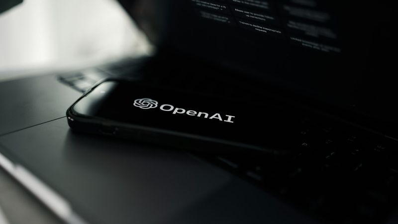 The image shows a smartphone displaying the OpenAI logo placed on a laptop keyboard. The background is muted, emphasizing the contrast between the black phone and the metallic laptop surface.