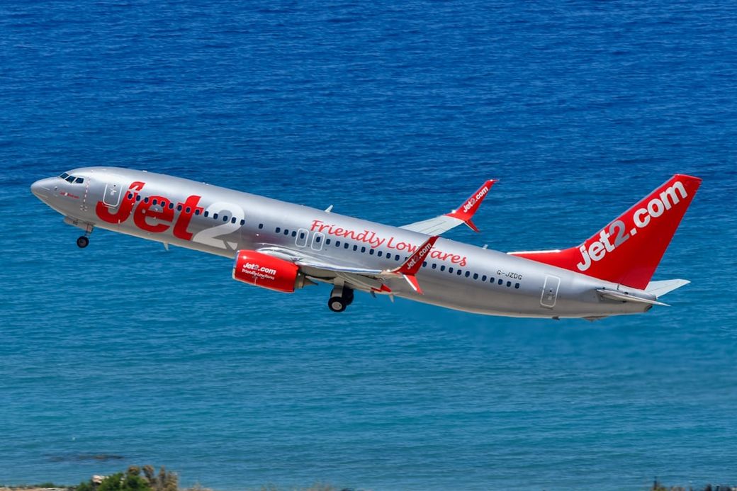 A Jet2.com airplane is taking off against a backdrop of blue sea. The aircraft is shown climbing into the sky with its distinctive branding visible on the fuselage. — Credit: Photo by David Syphers on Unsplash