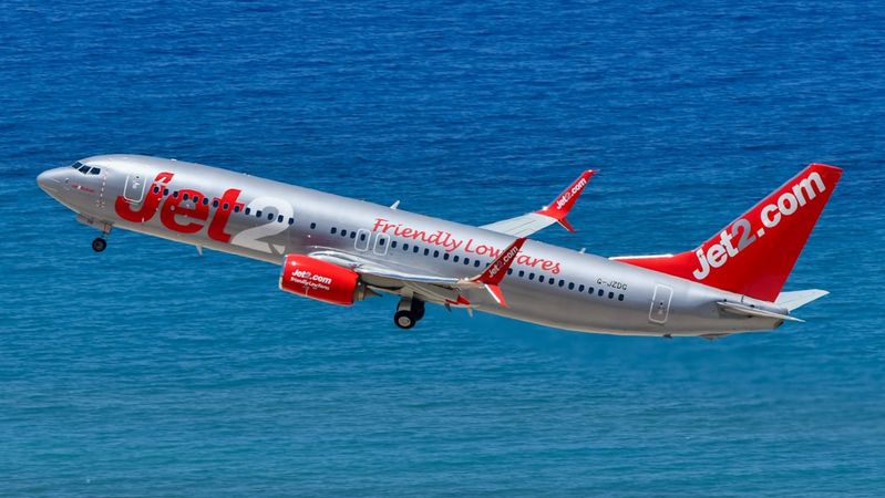 A Jet2.com airplane is taking off against a backdrop of blue sea. The aircraft is shown climbing into the sky with its distinctive branding visible on the fuselage.