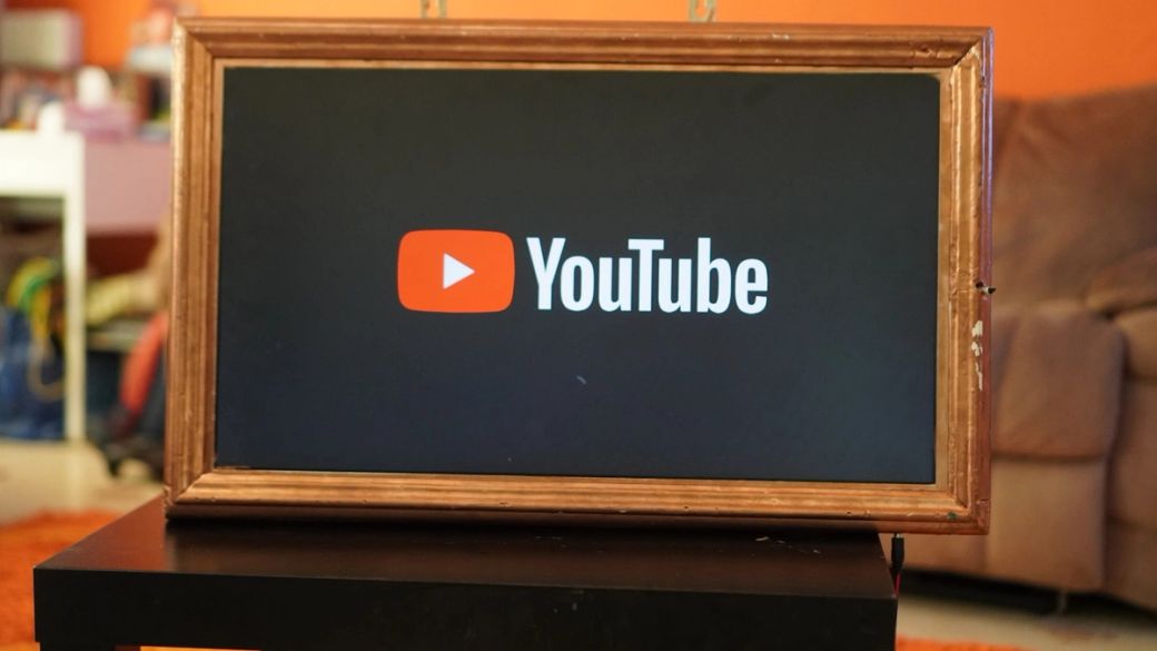 An old-fashioned framed television displays the YouTube logo prominently on a black screen. The background is orange, contrasting with the muted colors of the furniture in the scene. — Credit: Photo by حامد طه / Unsplash