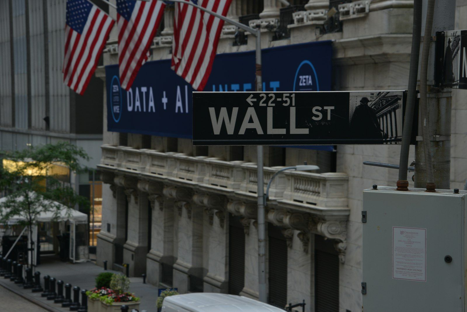 The image captures a street view of Wall Street, showcasing the iconic Wall Street sign beneath American flags. The backdrop features a large advertisement displaying 'DATA + AI' and logos of associated brands.