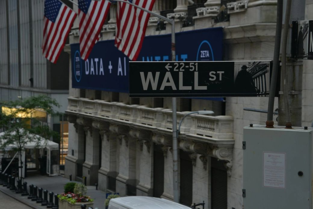 The image captures a street view of Wall Street, showcasing the iconic Wall Street sign beneath American flags. The backdrop features a large advertisement displaying 'DATA + AI' and logos of associated brands. — Credit: Photo by Robb Miller / Unsplash