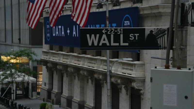 The image captures a street view of Wall Street, showcasing the iconic Wall Street sign beneath American flags. The backdrop features a large advertisement displaying 'DATA + AI' and logos of associated brands.