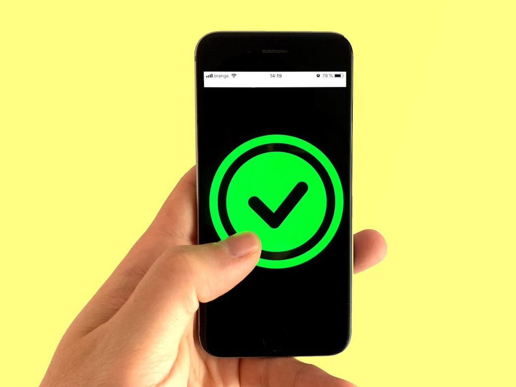 A person holds a smartphone displaying a glowing green checkmark icon on the screen. The background is a solid yellow color, creating a vibrant contrast that emphasizes the device and the icon. — Credit: Photo by Franck on Unsplash