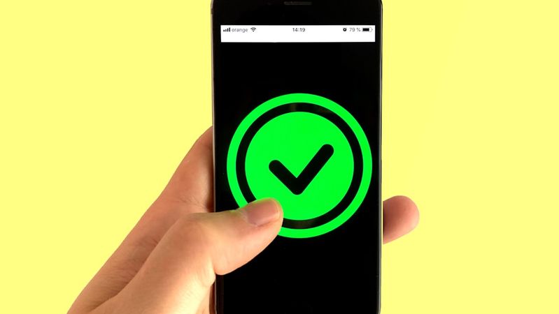 A person holds a smartphone displaying a glowing green checkmark icon on the screen. The background is a solid yellow color, creating a vibrant contrast that emphasizes the device and the icon.