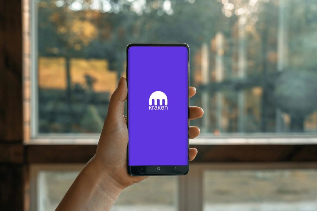 A person holding a smartphone displaying the Kraken cryptocurrency exchange app on its screen. The background features a blurred view of a window and nature, suggesting a relaxed environment. — Credit: Photo by PiggyBank / Unsplash