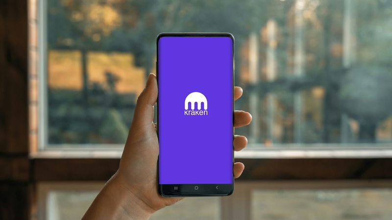 A person holding a smartphone displaying the Kraken cryptocurrency exchange app on its screen. The background features a blurred view of a window and nature, suggesting a relaxed environment.
