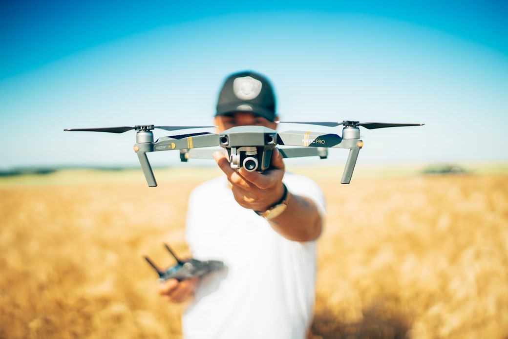 A person in a cap holds a drone in front of them, showcasing the device against a backdrop of a golden field under a clear blue sky. The individual appears ready to operate the drone, which is an example of modern technology in an agricultural setting. — Credit: Photo by david henrichs on Unsplash