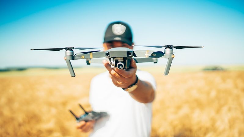 A person in a cap holds a drone in front of them, showcasing the device against a backdrop of a golden field under a clear blue sky. The individual appears ready to operate the drone, which is an example of modern technology in an agricultural setting.