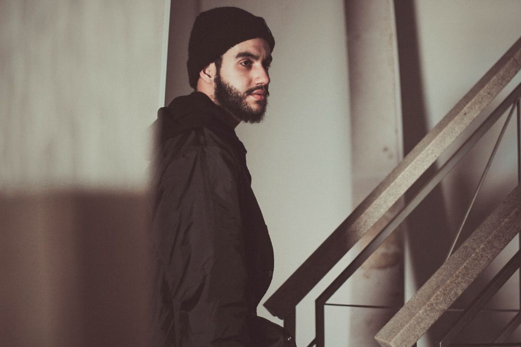 A young man with a beard and a beanie is seen standing on a staircase, looking off to the side. The image captures a moody atmosphere with shadowy lighting that emphasizes the man's facial features. — Credit: Photo by Allef Vinicius on Unsplash