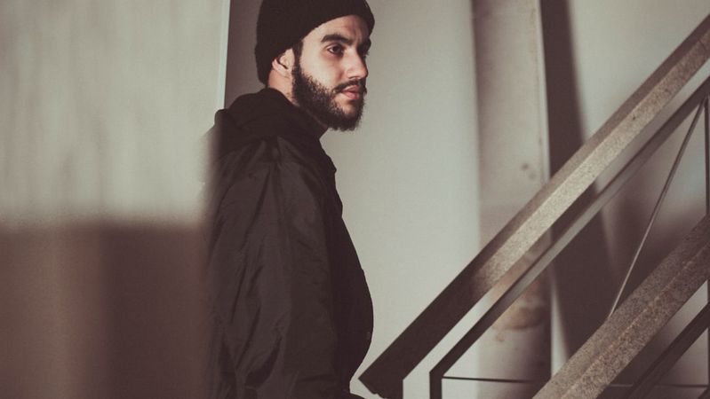 A young man with a beard and a beanie is seen standing on a staircase, looking off to the side. The image captures a moody atmosphere with shadowy lighting that emphasizes the man's facial features.