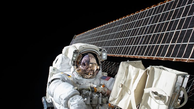 An astronaut in a space suit performs a spacewalk outside the International Space Station, with solar panels visible in the background. The astronaut's visor reflects the Earth and space, highlighting the vastness of the environment.
