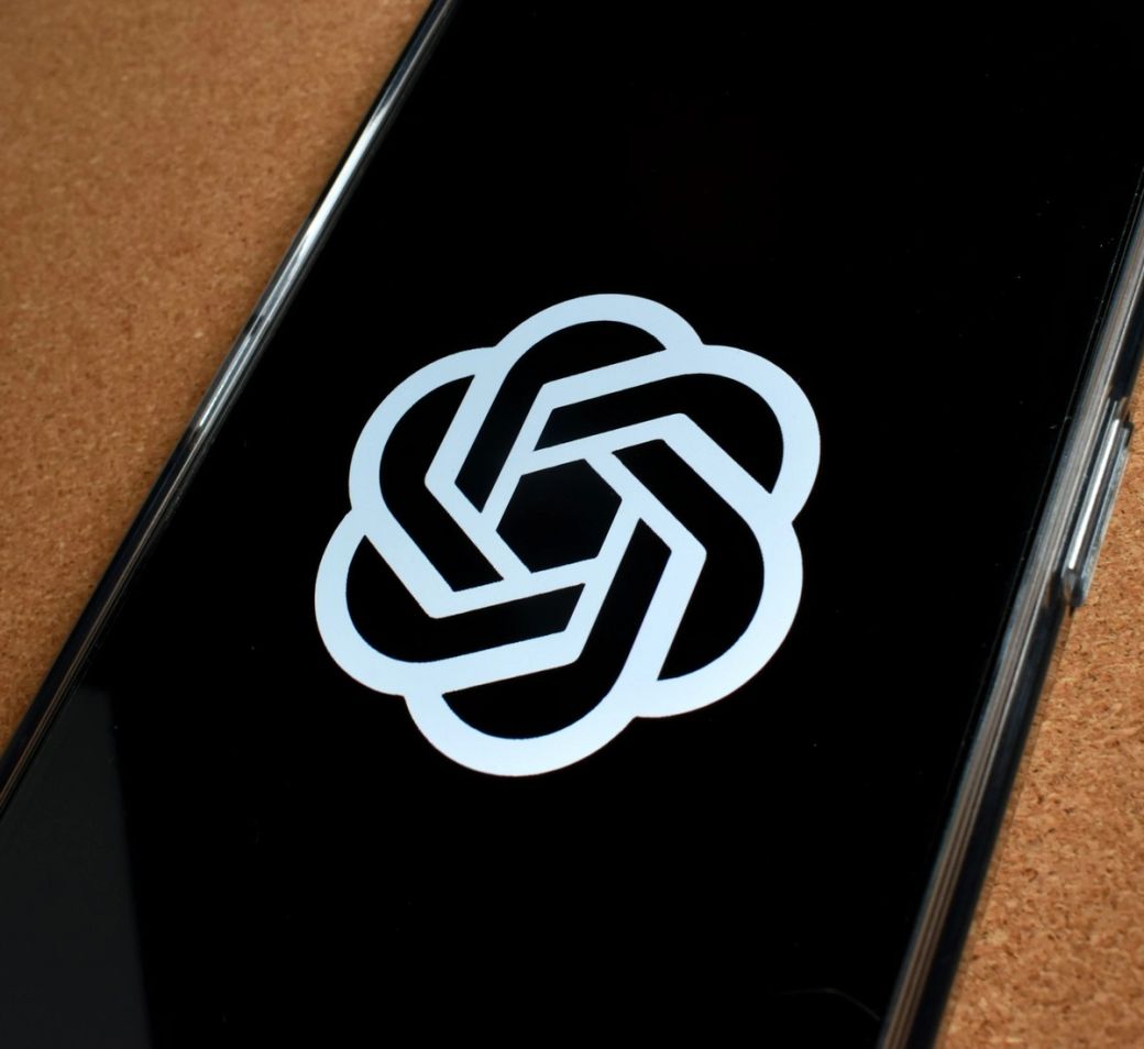 The image shows a close-up of a smartphone screen displaying the logo of OpenAI, which features a stylized, interwoven design. The background is black, emphasizing the logo's silver color. — Credit: Photo by Gavin Phillips / Unsplash