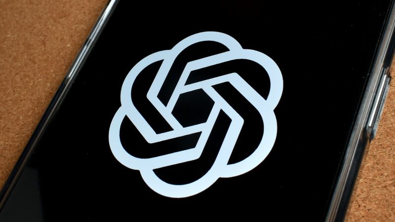 The image shows a close-up of a smartphone screen displaying the logo of OpenAI, which features a stylized, interwoven design. The background is black, emphasizing the logo's silver color.