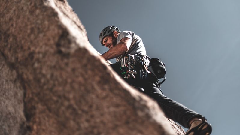 A climber is ascending a rocky cliff, showcasing the intensity and focus required for the sport. The image captures the climber from an elevated angle, emphasizing the challenging terrain and the climber's gear.