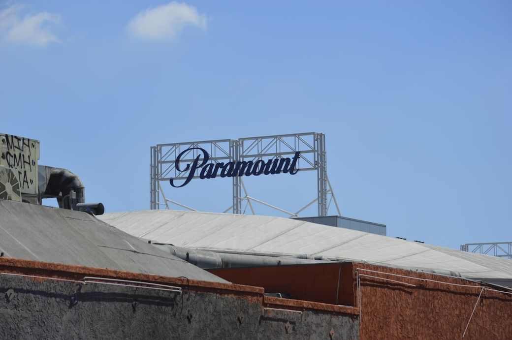 The image shows a clear blue sky above a building with a prominent sign that reads 'Paramount'. The sign is situated atop a structure with a sloped roof, typical of entertainment industry facilities. — Credit: Photo by Romain Malaunay on Unsplash