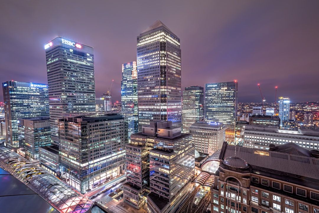 A panoramic view of a modern urban skyline at night, showcasing tall skyscrapers illuminated by city lights. Prominent buildings include those housing financial institutions, reflecting the vibrant economic activity of the area.
