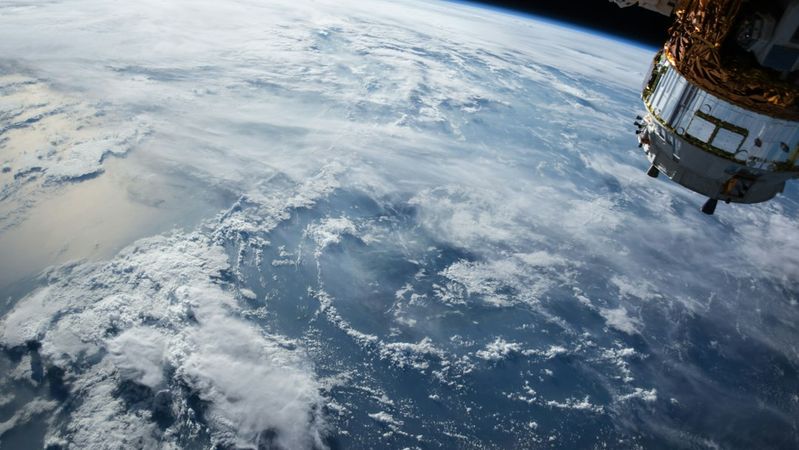 The image captures a view of Earth from space, showcasing clouds and the planet's surface against the backdrop of the vast atmosphere. A spacecraft component is visible in the foreground, highlighting the technology used in space exploration.
