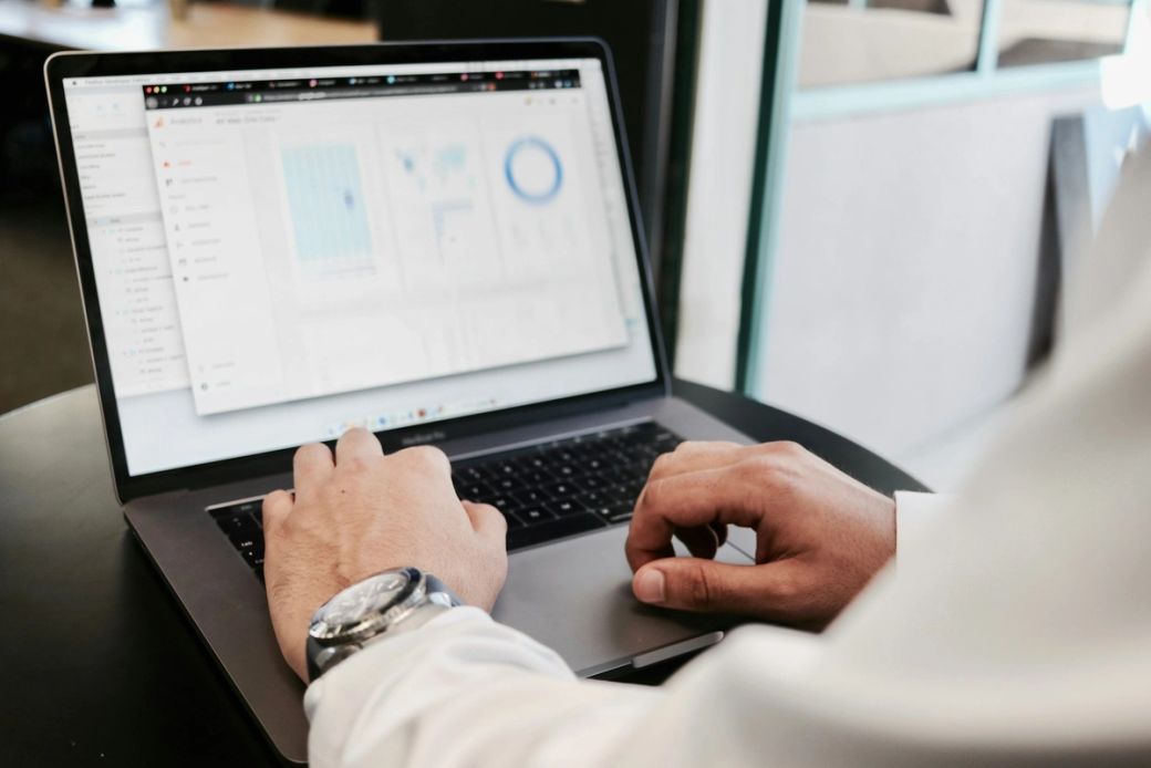 A close-up view of a person's hands working on a laptop, displaying various data charts and graphs on the screen. The setting appears to be an office or a workspace, emphasizing a focus on data analysis. — Credit: Photo by Campaign Creators / Unsplash