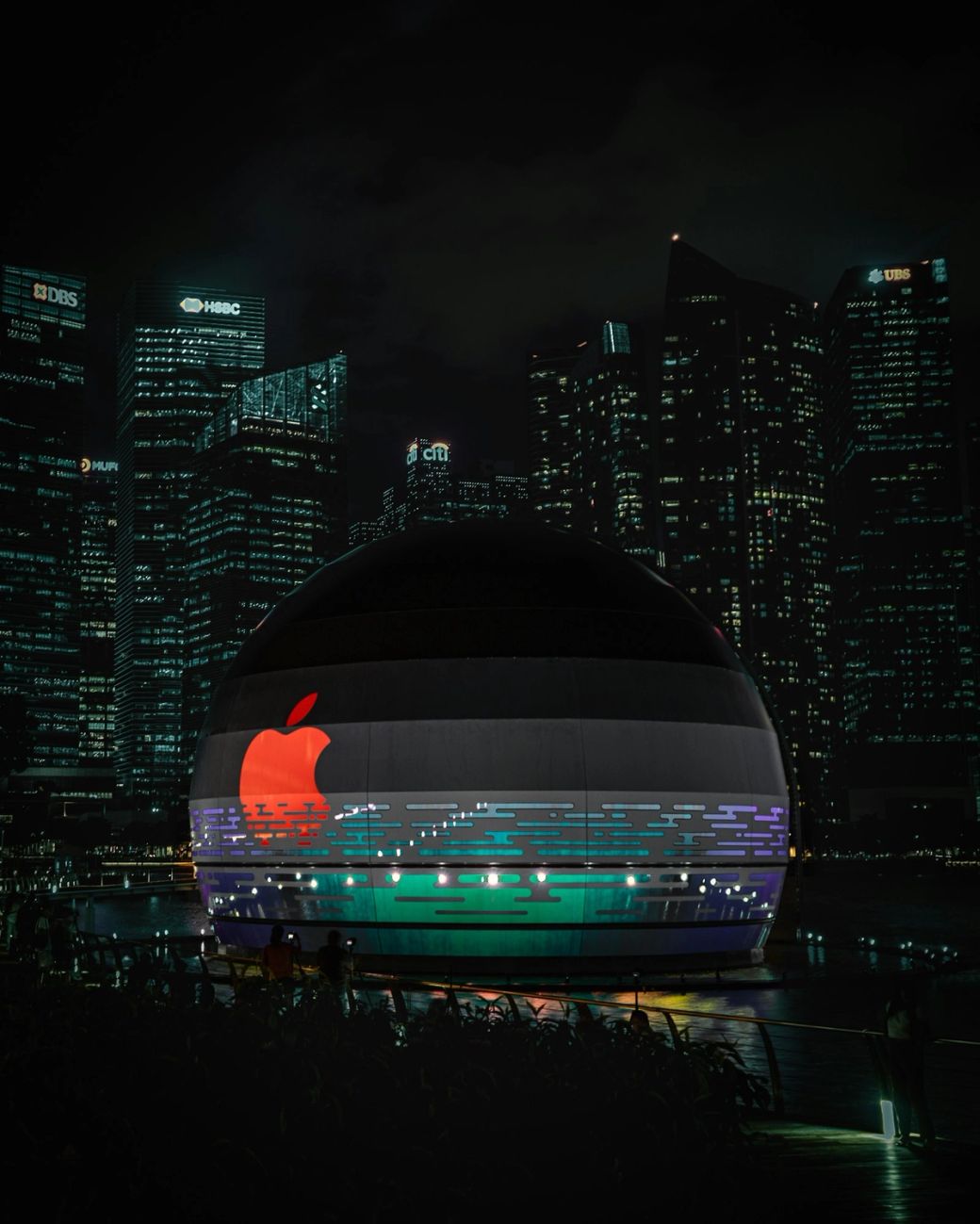The image showcases a circular structure illuminated with the Apple logo, set against a backdrop of a modern city skyline adorned with numerous skyscrapers. The scene is captured at night, emphasizing the vibrant city lights and the prominence of the Apple branding. — Credit: Photo by Bryan Low / Unsplash