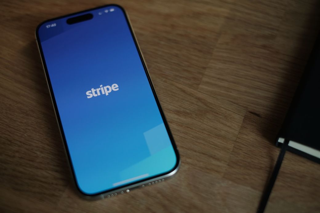 The image shows a smartphone displaying the Stripe app interface on its screen, with a blue gradient background. The device is placed on a wooden surface, alongside a journal or notebook. — Credit: Photo by appshunter.io on Unsplash