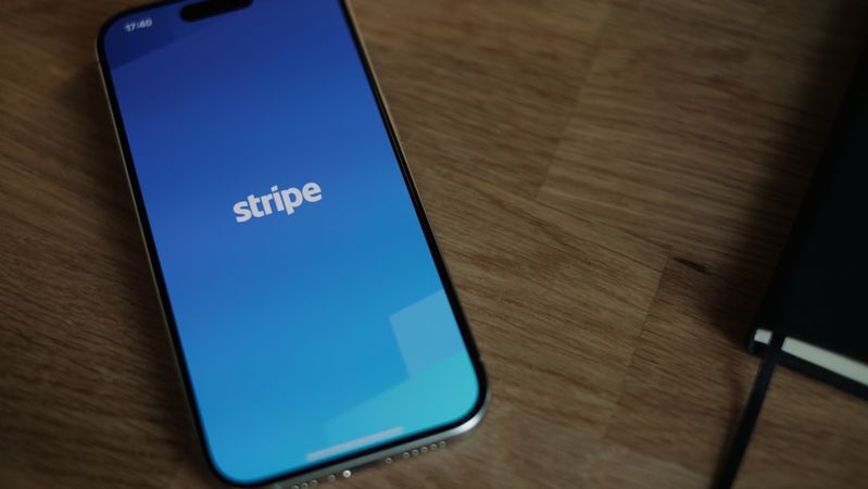 The image shows a smartphone displaying the Stripe app interface on its screen, with a blue gradient background. The device is placed on a wooden surface, alongside a journal or notebook.