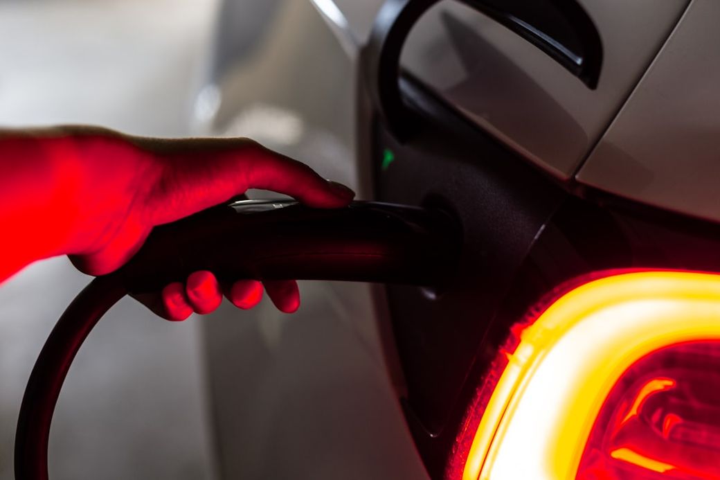 An electric vehicle is being charged, with a hand holding the charging connector. The vehicle's illuminated rear light is visible, highlighting its modern design. — Credit: Photo by Priscilla Du Preez 🇨🇦 on Unsplash