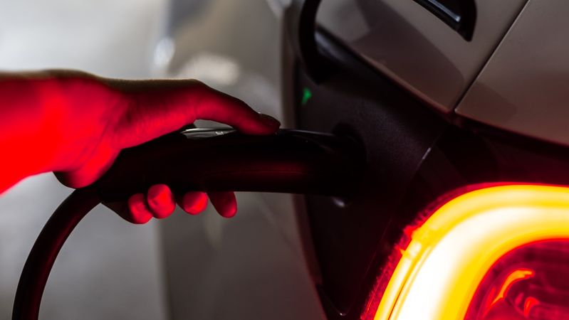 An electric vehicle is being charged, with a hand holding the charging connector. The vehicle's illuminated rear light is visible, highlighting its modern design.