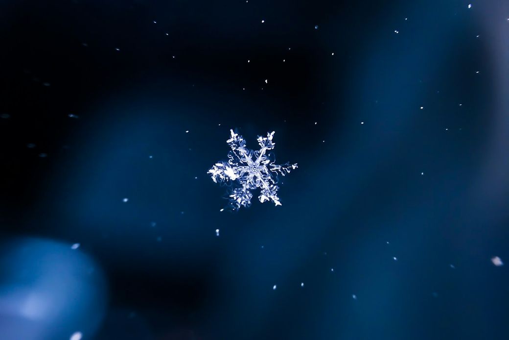 A close-up view of a single snowflake captured against a blue background. The intricate patterns of the snowflake are highlighted in sharp detail, showcasing the unique structure of the ice crystals. — Credit: Photo by Darius Cotoi / Unsplash
