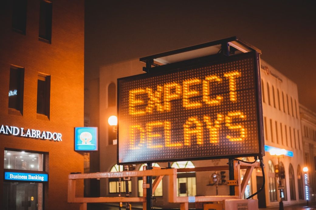 A digital sign displaying the message 'EXPECT DELAYS' is prominently featured in an urban environment during low visibility conditions. The background includes a bank and a brick building, indicating a commercial area. — Credit: Photo by Erik Mclean on Unsplash