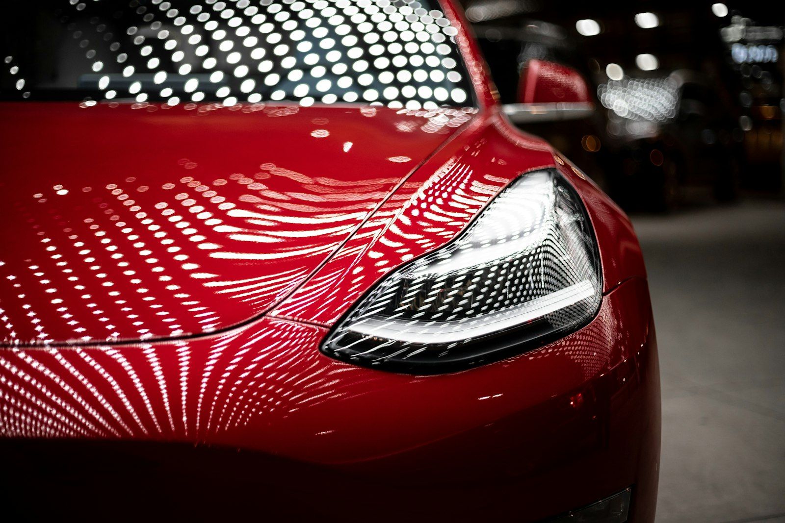 The image features a close-up view of a glossy red car, highlighting its sleek design and distinctive headlight features. The reflections of numerous lights create a dynamic and vibrant background, emphasizing the car's modern aesthetics.