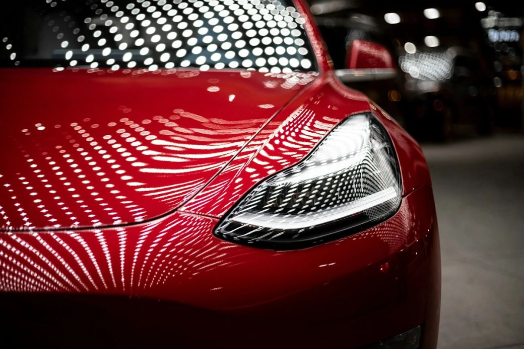 The image features a close-up view of a glossy red car, highlighting its sleek design and distinctive headlight features. The reflections of numerous lights create a dynamic and vibrant background, emphasizing the car's modern aesthetics. — Credit: Photo by Vlad Tchompalov / Unsplash