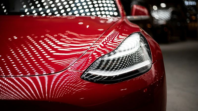 The image features a close-up view of a glossy red car, highlighting its sleek design and distinctive headlight features. The reflections of numerous lights create a dynamic and vibrant background, emphasizing the car's modern aesthetics.