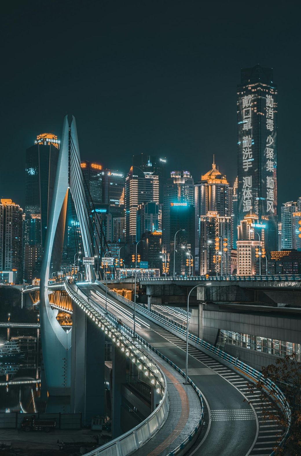 The image depicts a modern bridge illuminated at night, leading towards a vibrant city skyline filled with skyscrapers. The urban landscape showcases a blend of contemporary architecture and bright advertisements. — Credit: Photo by Jerry Wang on Unsplash