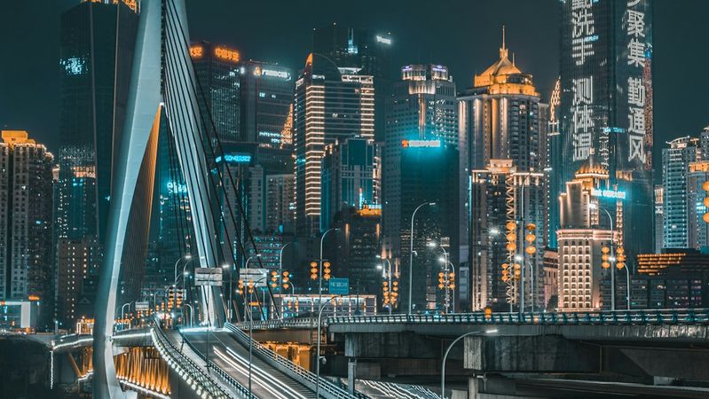 The image depicts a modern bridge illuminated at night, leading towards a vibrant city skyline filled with skyscrapers. The urban landscape showcases a blend of contemporary architecture and bright advertisements.