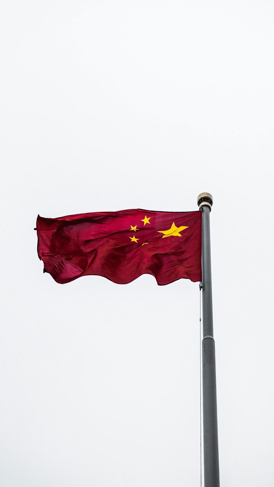 The image features the national flag of China fluttering on a flagpole against a cloudy sky. The flag is predominantly red with five yellow stars, arranged in a specific formation. — Credit: Photo by Alejandro Luengo on Unsplash