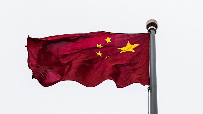 The image features the national flag of China fluttering on a flagpole against a cloudy sky. The flag is predominantly red with five yellow stars, arranged in a specific formation.