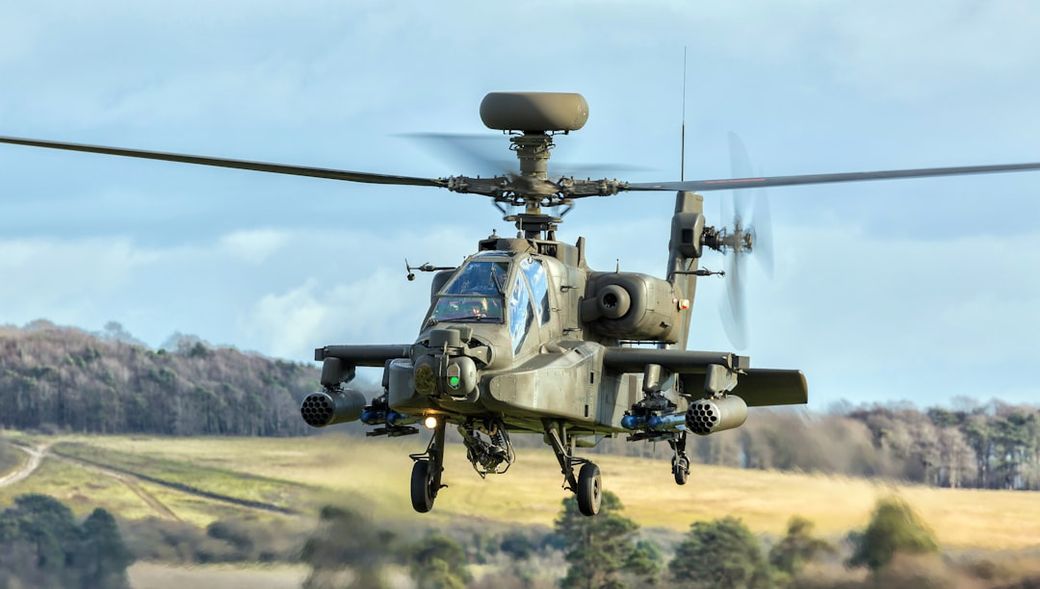 An attack helicopter is captured mid-flight against a backdrop of a cloudy sky and grassy terrain. The helicopter displays military features and is equipped with visible armaments. — Credit: Photo by DON JACKSON-WYATT on Unsplash