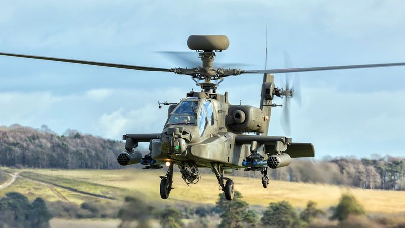 An attack helicopter is captured mid-flight against a backdrop of a cloudy sky and grassy terrain. The helicopter displays military features and is equipped with visible armaments.