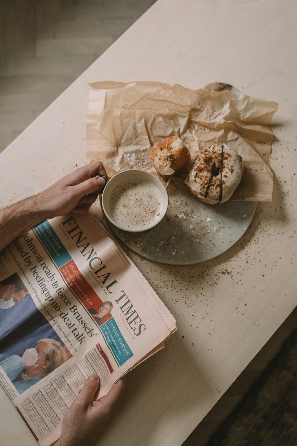 A person is sitting at a table holding a cup of coffee while reading the Financial Times newspaper. The table is set with a plate containing pastries, alongside scattered crumbs. — Credit: Photo by Toa Heftiba on Unsplash