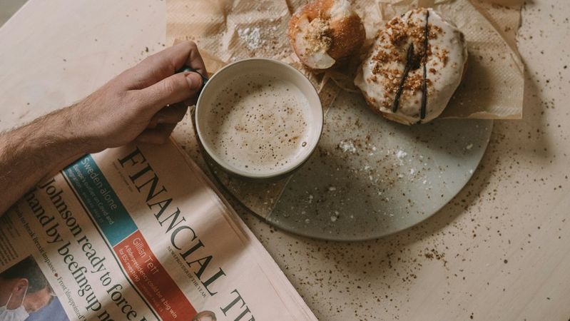 A person is sitting at a table holding a cup of coffee while reading the Financial Times newspaper. The table is set with a plate containing pastries, alongside scattered crumbs.