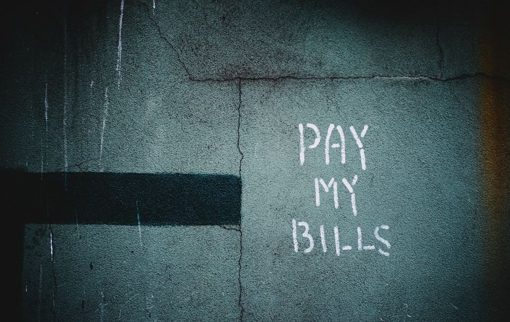 The image features graffiti on a textured wall, predominantly showcasing the phrase 'PAY MY BILLS' in white paint. The background has a dark blue-green hue, adding to the urban aesthetic. — Credit: Photo by Hiroshi Kimura on Unsplash