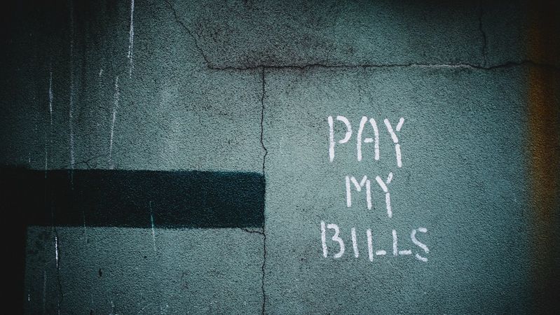 The image features graffiti on a textured wall, predominantly showcasing the phrase 'PAY MY BILLS' in white paint. The background has a dark blue-green hue, adding to the urban aesthetic.