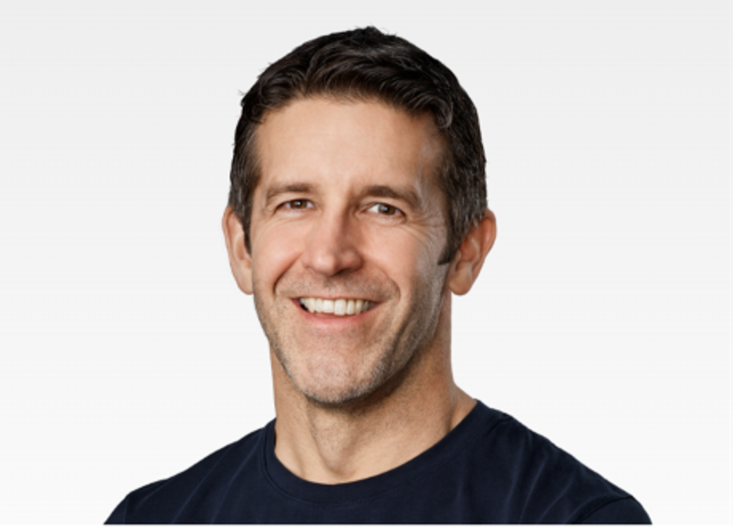 The image features a close-up portrait of a middle-aged man with short dark hair and a friendly smile. He is wearing a dark shirt against a light gray background, projecting a professional and approachable demeanor.