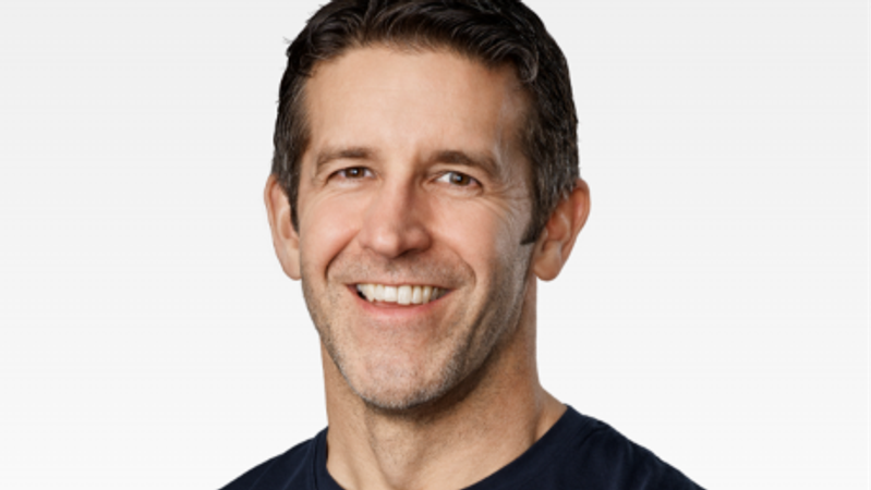 The image features a close-up portrait of a middle-aged man with short dark hair and a friendly smile. He is wearing a dark shirt against a light gray background, projecting a professional and approachable demeanor.