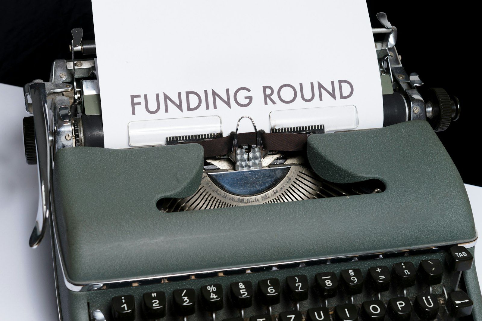 The image features a vintage typewriter with a sheet of paper inserted, displaying the text 'FUNDING ROUND'. The composition highlights the typewriter, emphasizing the concept of fundraising or investment.