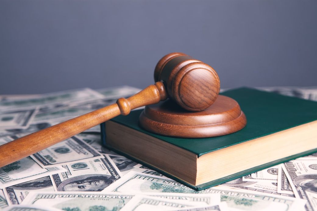 The image features a wooden gavel resting on top of a closed green legal book, with U.S. dollar bills scattered in the background. This composition visually represents the intersection of law and finance. — Credit: Photo by Sasun Bughdaryan on Unsplash