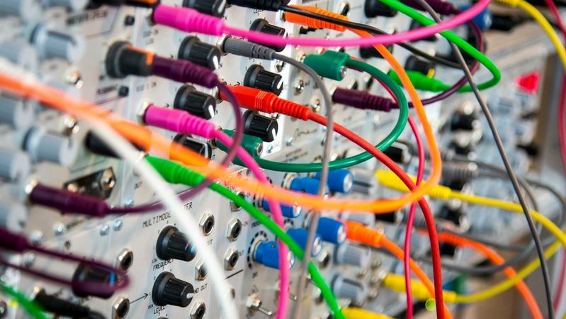 The image shows a close-up of an electronic device with numerous colorful cables and wires connected to various ports and knobs. This setup is typical in sound synthesis, demonstrating the complexity of modular synthesizers.